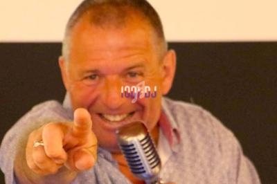 Homme souriant pointant vers l'objectif, tenant un microphone devant un fond neutre.