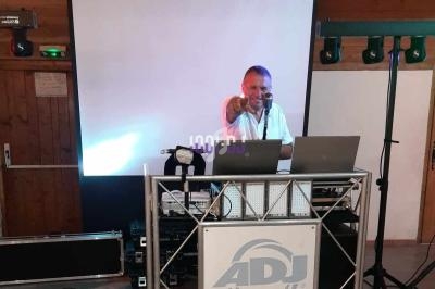 Un DJ souriant anime une soirée depuis une table de mixage avec un écran blanc en arrière-plan.