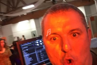 Homme s'exprimant devant un micro, avec un ordinateur portable affichant une playlist en arrière-plan dans une salle.