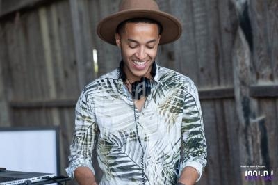 Un DJ souriant portant un chapeau et un casque audio, debout devant un mur en bois.