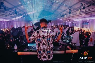 Un DJ anime une soirée dans une salle éclairée par des jeux de lumière, avec un public dansant en arrière-plan.