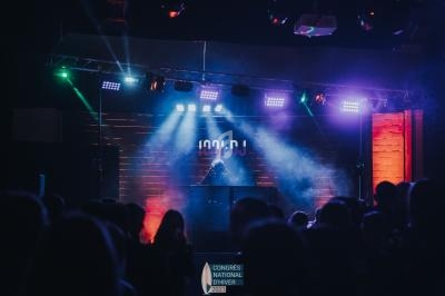 Un DJ mixe sur une scène éclairée par des projecteurs colorés, entouré d'une foule dans une ambiance de concert.