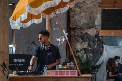 Un DJ mixe de la musique sous un parasol jaune dans un décor industriel avec des éléments en bois.