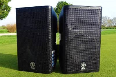 Deux enceintes noires posées sur une pelouse verte, avec des arbres et un ciel dégagé en arrière-plan.