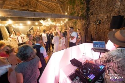 Groupe de personnes dansant sous des guirlandes lumineuses dans une salle en bois, avec un DJ en premier plan.