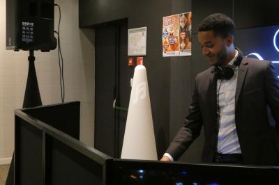 Un DJ en costume mixe de la musique derrière une table dans un espace intérieur avec des affiches au mur.