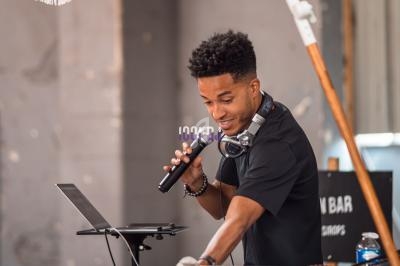 Un DJ mixe de la musique sur une table équipée sous un parasol, tenant un micro dans un cadre urbain.