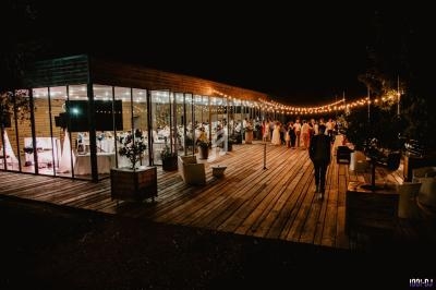 Salle de réception vitrée éclairée la nuit, avec des invités rassemblés sur une terrasse en bois décorée de guirlandes…