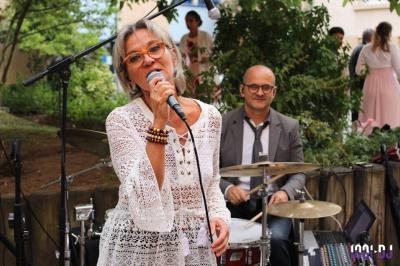 Une femme chante au micro devant un batteur, dans un cadre extérieur avec des arbres en arrière-plan.