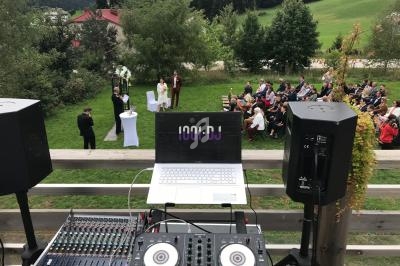 Vue depuis une table de mixage DJ sur une cérémonie en extérieur avec invités assis et paysage verdoyant en arrière-plan.