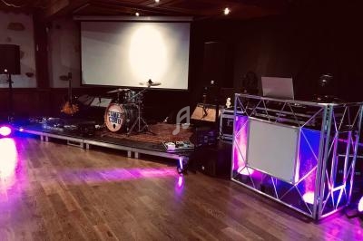Scène avec batterie, guitare, amplis et éclairages, à côté d'une régie DJ sur une piste en bois.