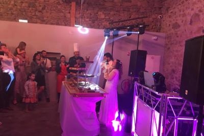 Un couple en tenue de mariage coupe un gâteau devant des invités dans une salle en pierre éclairée par des lumières colorées.