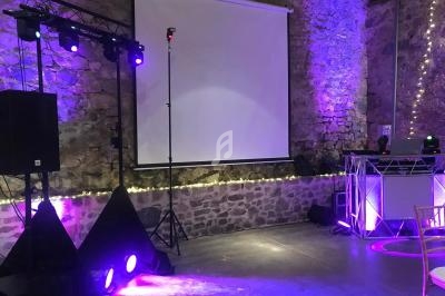 Salle en pierre décorée de guirlandes lumineuses avec écran de projection, éclairages violets et matériel de sonorisation.