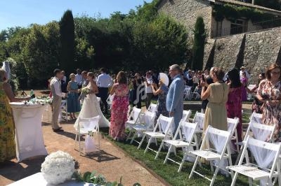 Cérémonie de mariage en extérieur avec des invités assis et debout, entourés de verdure et d'un bâtiment en pierre.