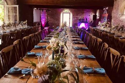 Grande salle en pierre avec de longues tables en bois dressées pour un repas, décorées de fleurs et de serviettes bleues.