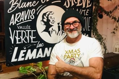 Homme souriant portant un bonnet et un t-shirt, posant devant une affiche illustrée mentionnant des lieux alpins.