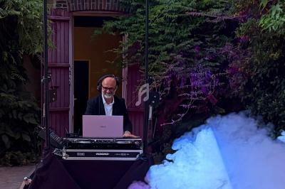 Un DJ mixe en plein air sous un éclairage coloré, entouré de fumée et de végétation.