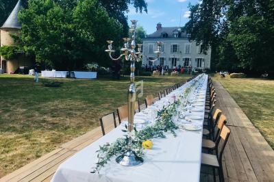Grande table dressée en extérieur avec nappes blanches, chandeliers et décoration végétale, devant un manoir.
