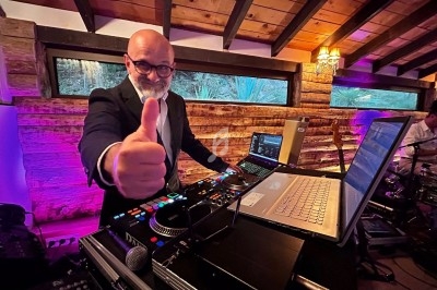 Un DJ souriant en costume fait un signe de pouce levé derrière une table de mixage dans une salle en bois éclairée.