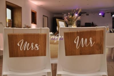 Chaises blanches décorées de panneaux en bois gravés ’Mrs’ et ’Mr’, dans une salle de réception éclairée.