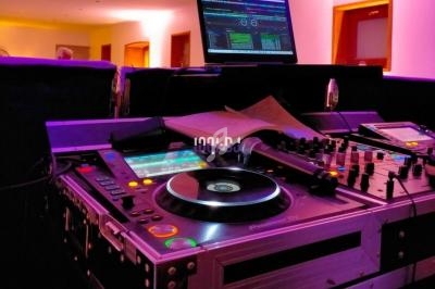 Table de mixage avec platines, écran d'ordinateur affichant un logiciel audio, dans une pièce éclairée de lumières colorées.
