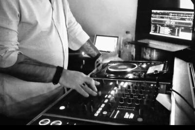 Un DJ en chemise blanche utilise une table de mixage avec des écrans d'ordinateur en arrière-plan.