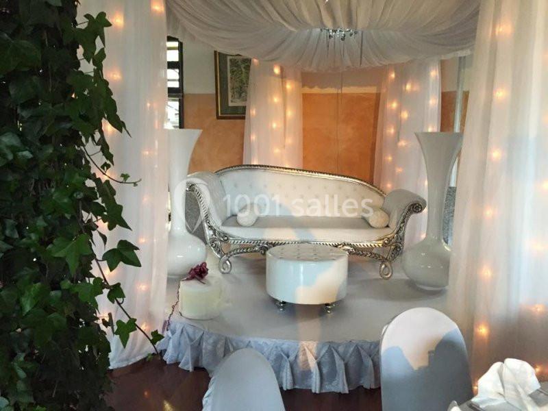 Canapé blanc orné dans un décor élégant avec rideaux, lumières tamisées et plantes vertes en premier plan.