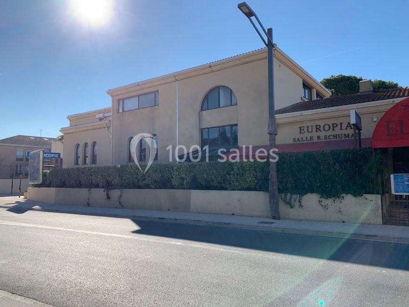 Bâtiment beige avec grandes fenêtres en arc, enseigne ’Europia Salle R. Schuman’, bordé d'une haie et situé en bord de route.