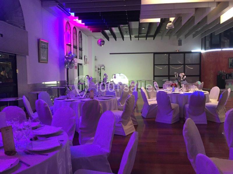 Salle de réception décorée avec des tables rondes dressées, chaises blanches et éclairage tamisé aux tons violets.