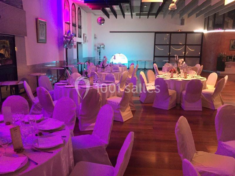 Salle de réception décorée avec des tables rondes dressées, chaises couvertes de housses blanches et éclairage tamisé.