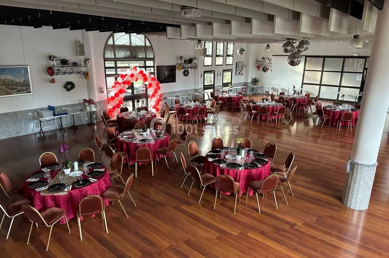 Salle de réception avec parquet, tables rondes dressées et arche décorative en ballons rouges et blancs.
