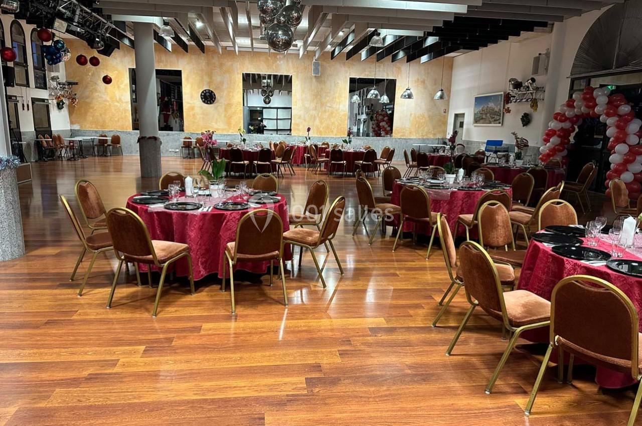 Salle de réception avec tables rondes dressées, nappes rouges, chaises marron et sol en parquet.