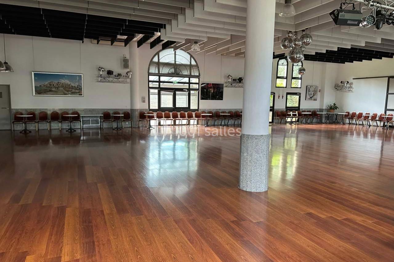 Salle spacieuse avec parquet en bois, chaises alignées le long des murs et grandes fenêtres laissant entrer la lumière.