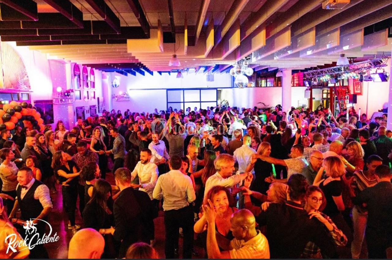 Des personnes dansent en groupe dans une grande salle éclairée par des lumières colorées, ambiance festive.