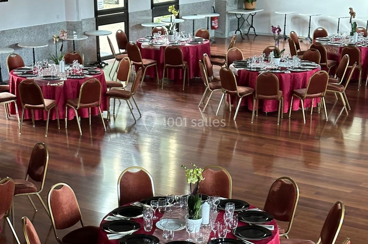 Salle de réception avec tables rondes dressées, nappes rouges, chaises marron et décoration florale.