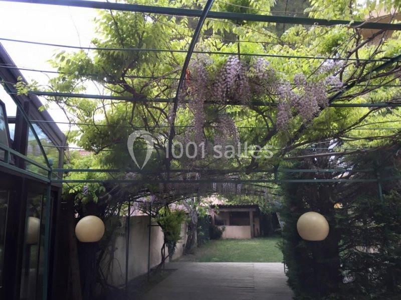 Pergola en métal recouverte de glycines fleuries, entourée de verdure, avec des lampes suspendues et un jardin en arrière…