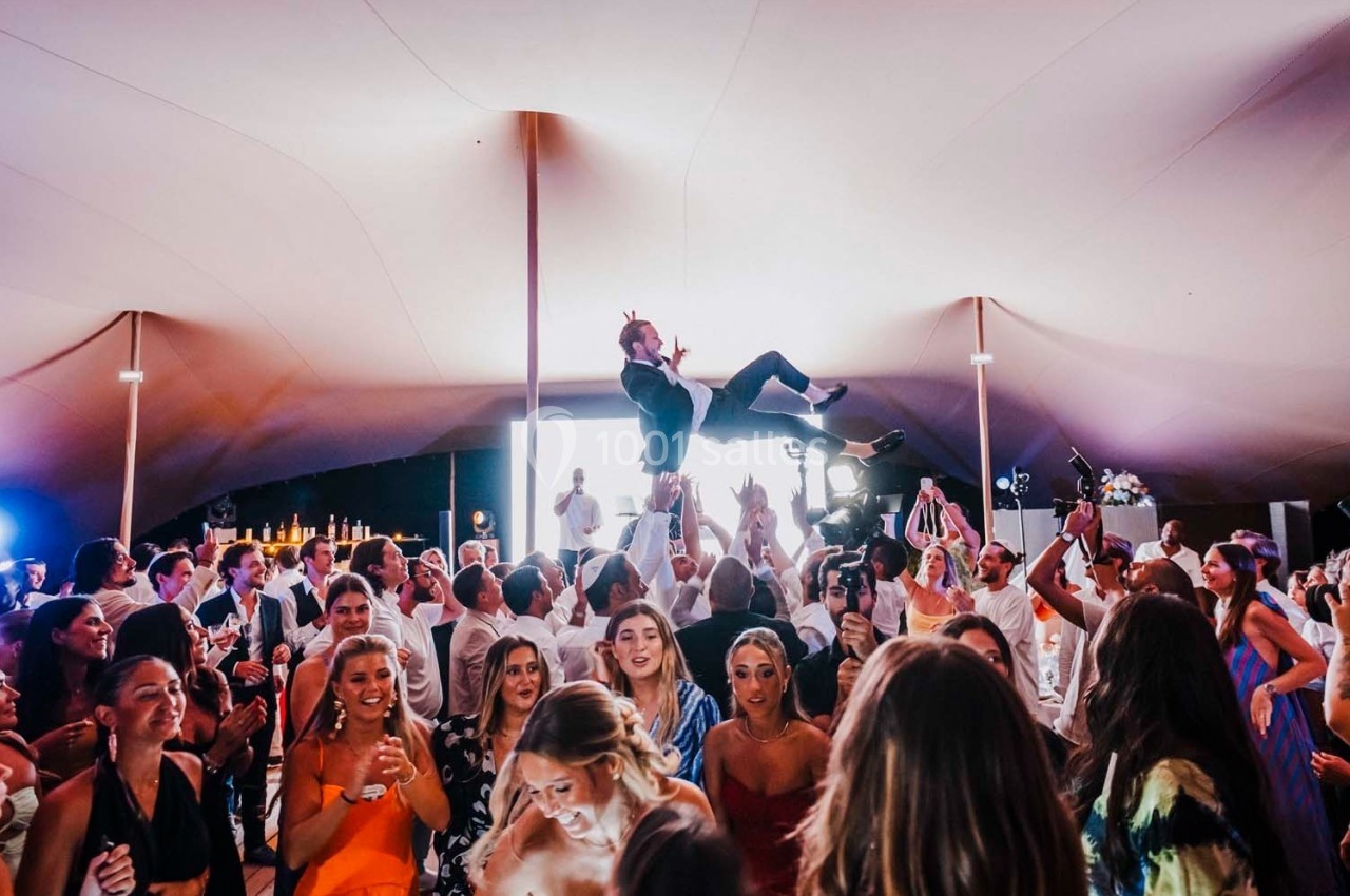 Personnes dansant sous une grande tente lors d'une fête, un homme est lancé en l'air par un groupe au centre.