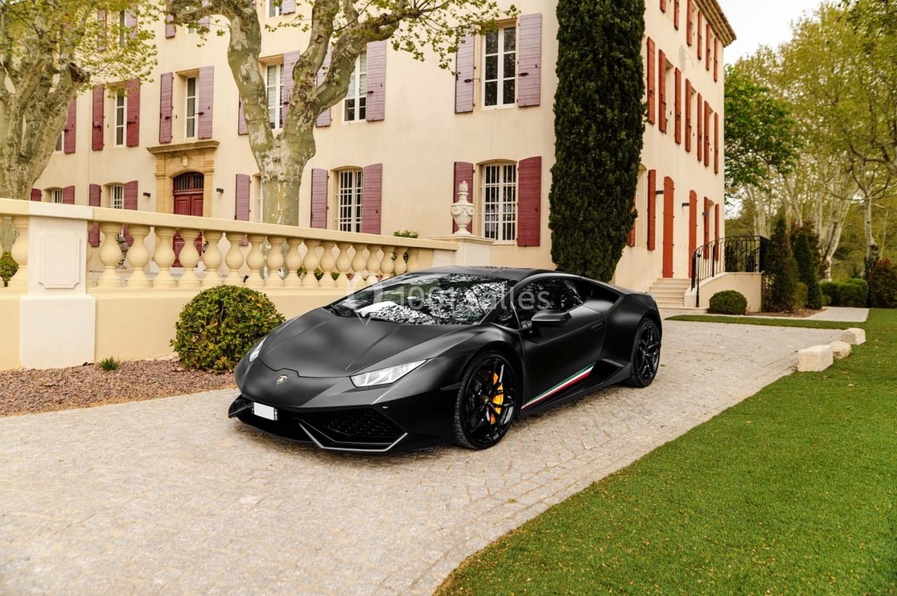 Voiture de sport noire garée sur une allée pavée devant une grande maison aux volets rouges entourée d'arbres.