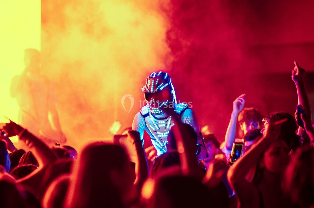 Un artiste en costume lumineux se produit sur scène entouré d'une foule dans une ambiance colorée et enfumée.
