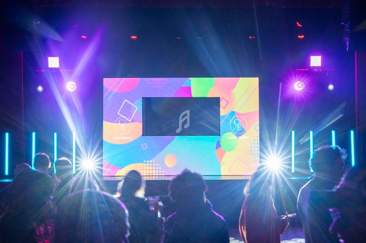 Scène illuminée avec écran coloré au centre, éclairages vifs et silhouettes de spectateurs au premier plan.