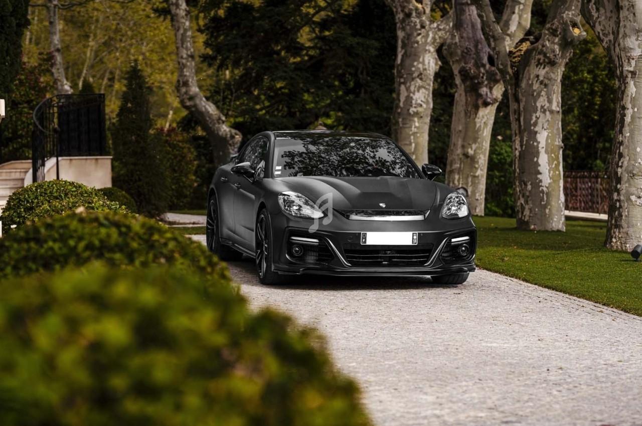 Voiture noire garée sur une allée pavée bordée de buissons taillés et d'arbres imposants.