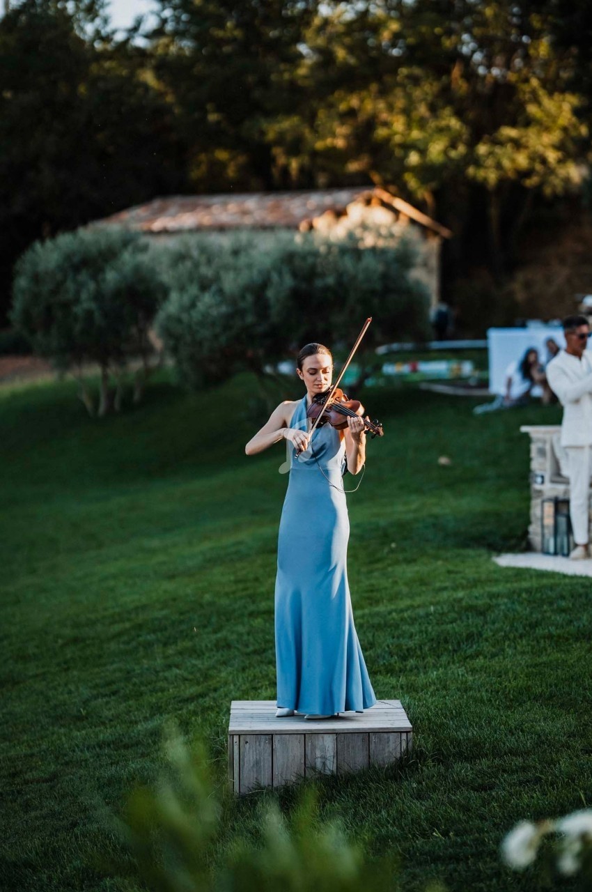 Une femme en robe bleue joue du violon sur une petite estrade en bois dans un jardin verdoyant.
