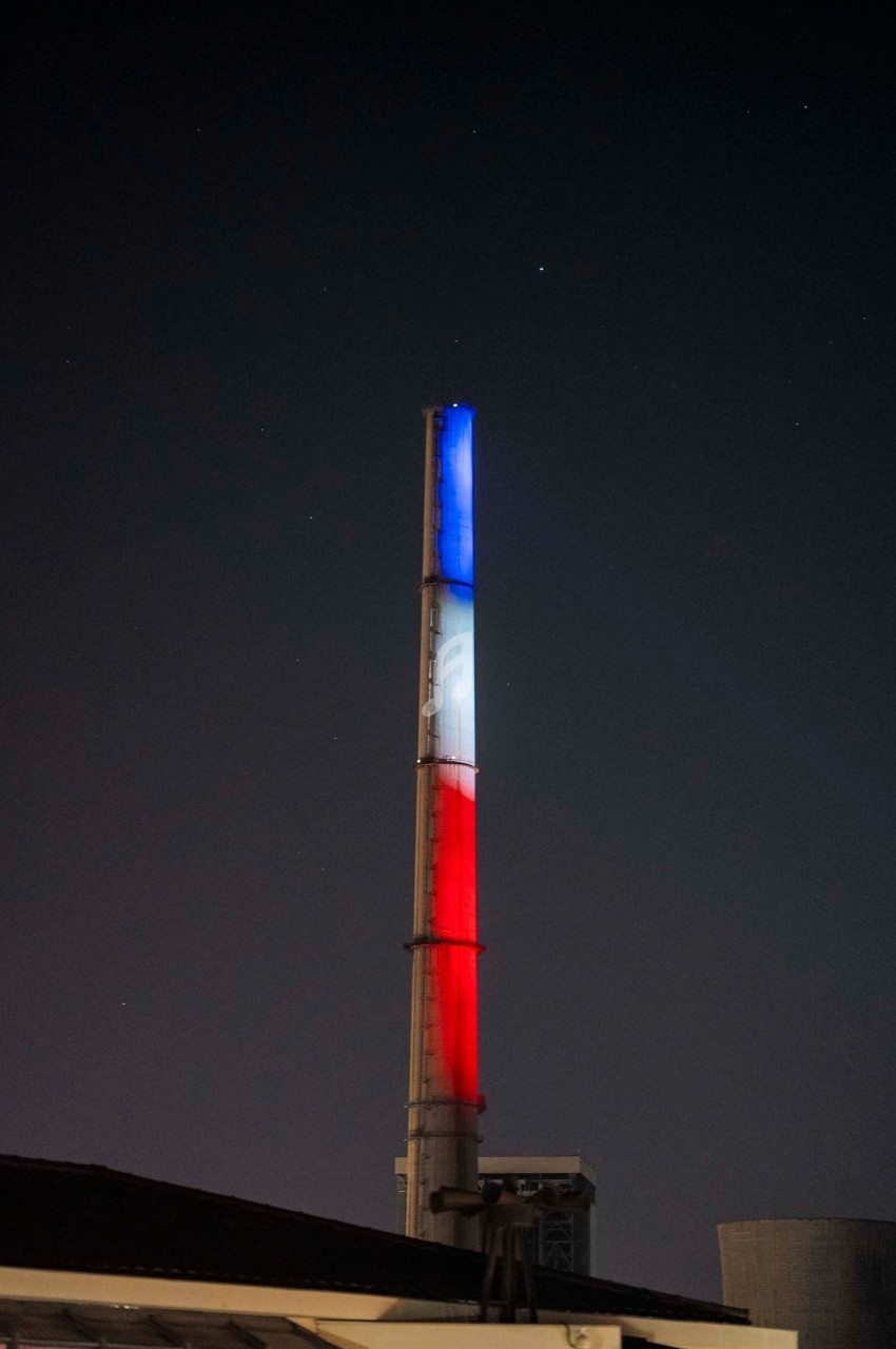 Cheminée industrielle illuminée en bleu, blanc et rouge dans un paysage nocturne.