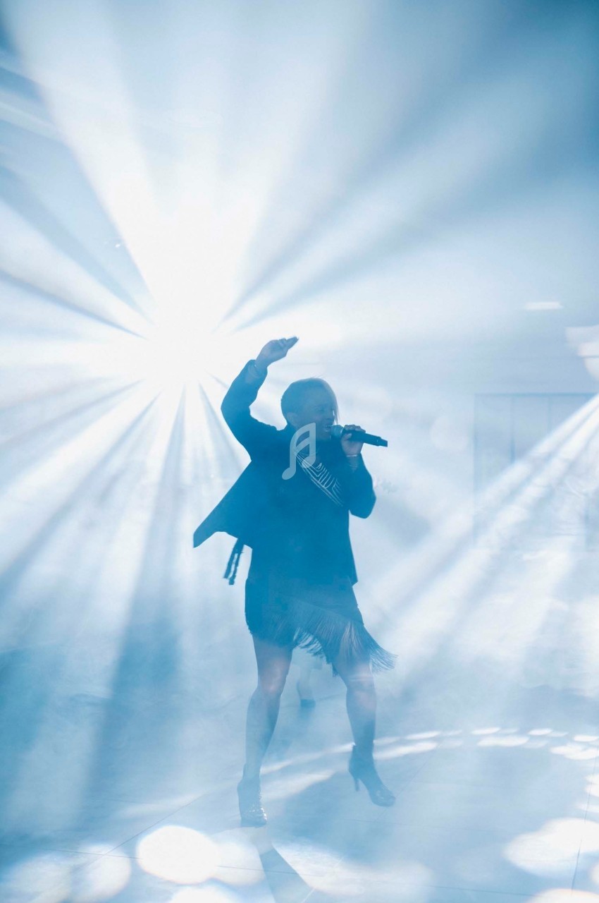 Une chanteuse sur scène, éclairée par des faisceaux lumineux dans une ambiance de fumée.