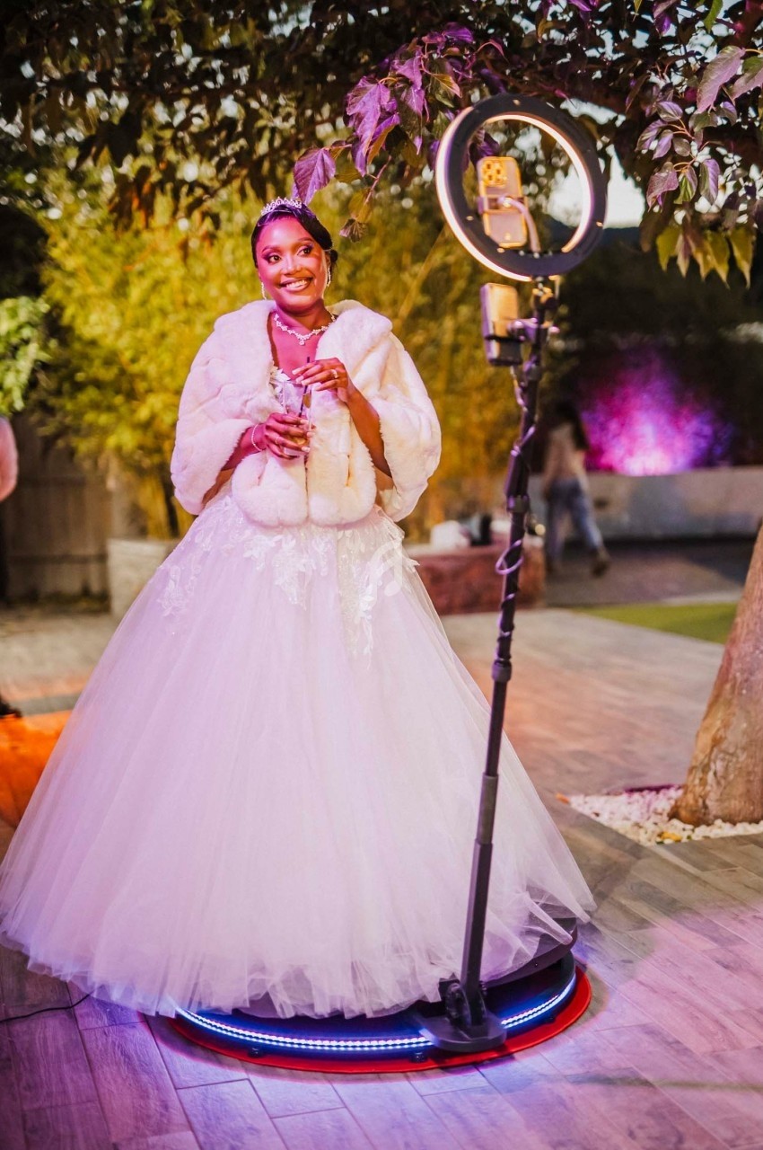 Une femme en robe de mariée blanche sourit, tenant un verre, devant un anneau lumineux dans un jardin éclairé.