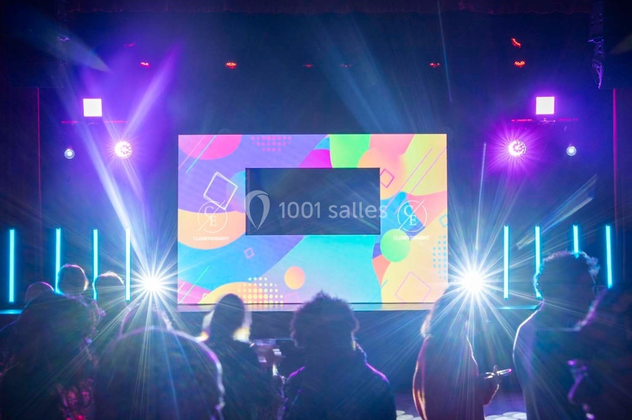 Scène illuminée avec écran coloré au centre, éclairages vifs et silhouettes de spectateurs au premier plan.