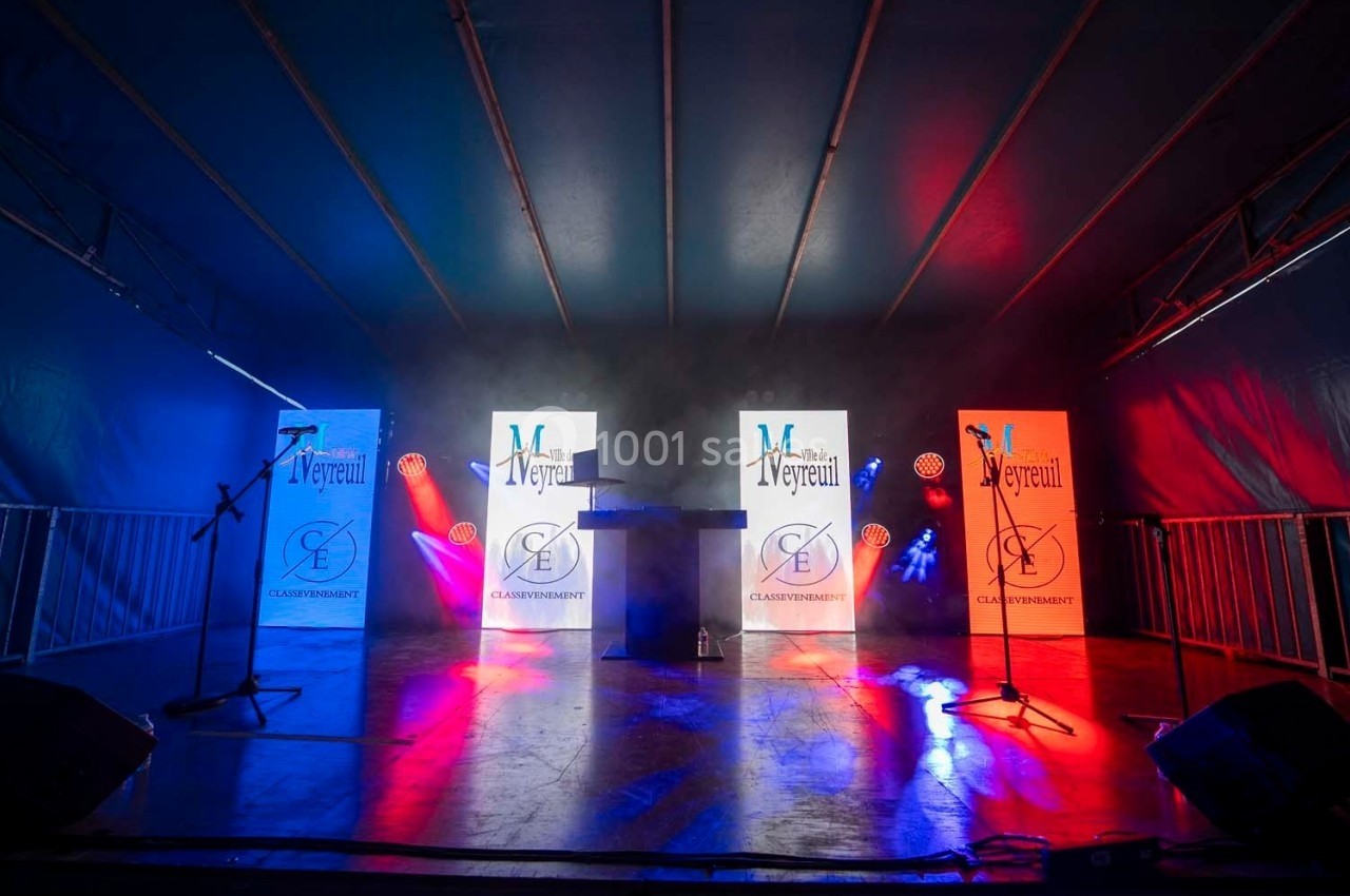 Scène éclairée en bleu, blanc et rouge avec deux micros, un pupitre central et des panneaux lumineux en arrière-plan.