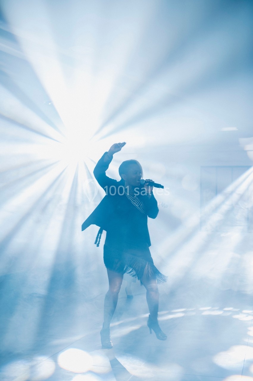 Une chanteuse sur scène, éclairée par des faisceaux lumineux dans une ambiance de fumée.