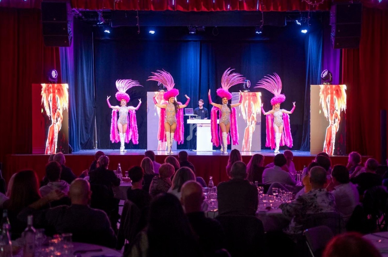 Des danseuses en costumes roses à plumes se produisent sur scène devant un public assis dans une salle éclairée.
