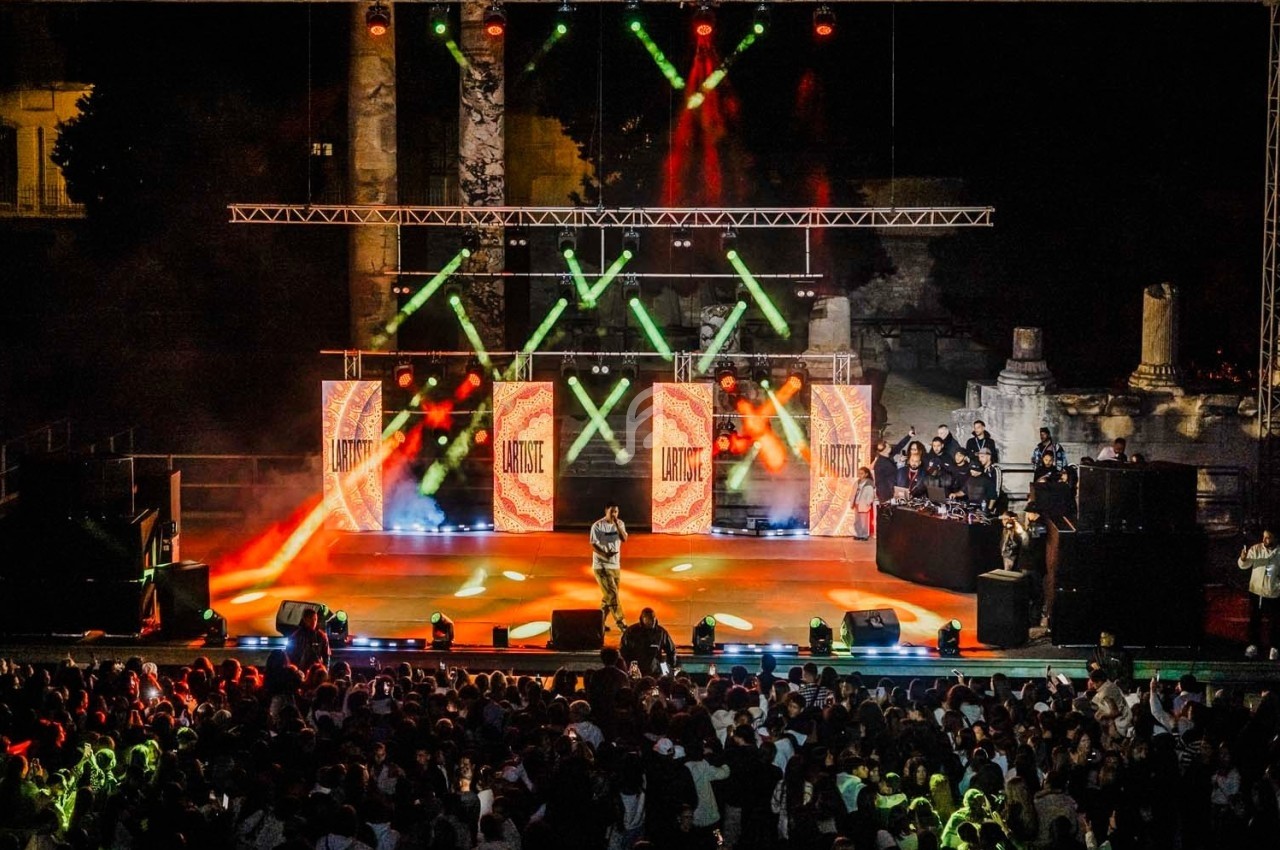 Un artiste chante sur une scène illuminée avec des écrans et des jeux de lumière, devant un public nombreux en plein air.